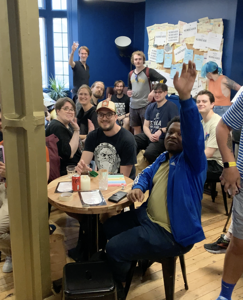 Group of people gathered in a cozy room, some sitting at a round wooden table with drinks and menus, others standing or sitting on chairs, with a blue wall and bulletin boards in the background.