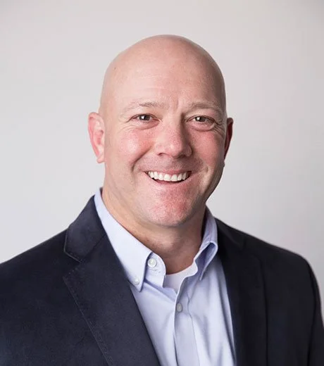 A smiling bald man in a dark suit jacket and light blue shirt against a plain light gray background.