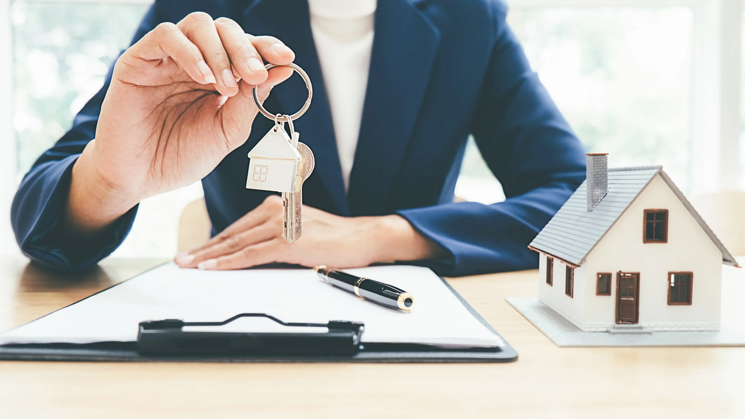 Person holding a key with a house-shaped keychain, sitting at a desk with a small model house and a clipboard with a pen.