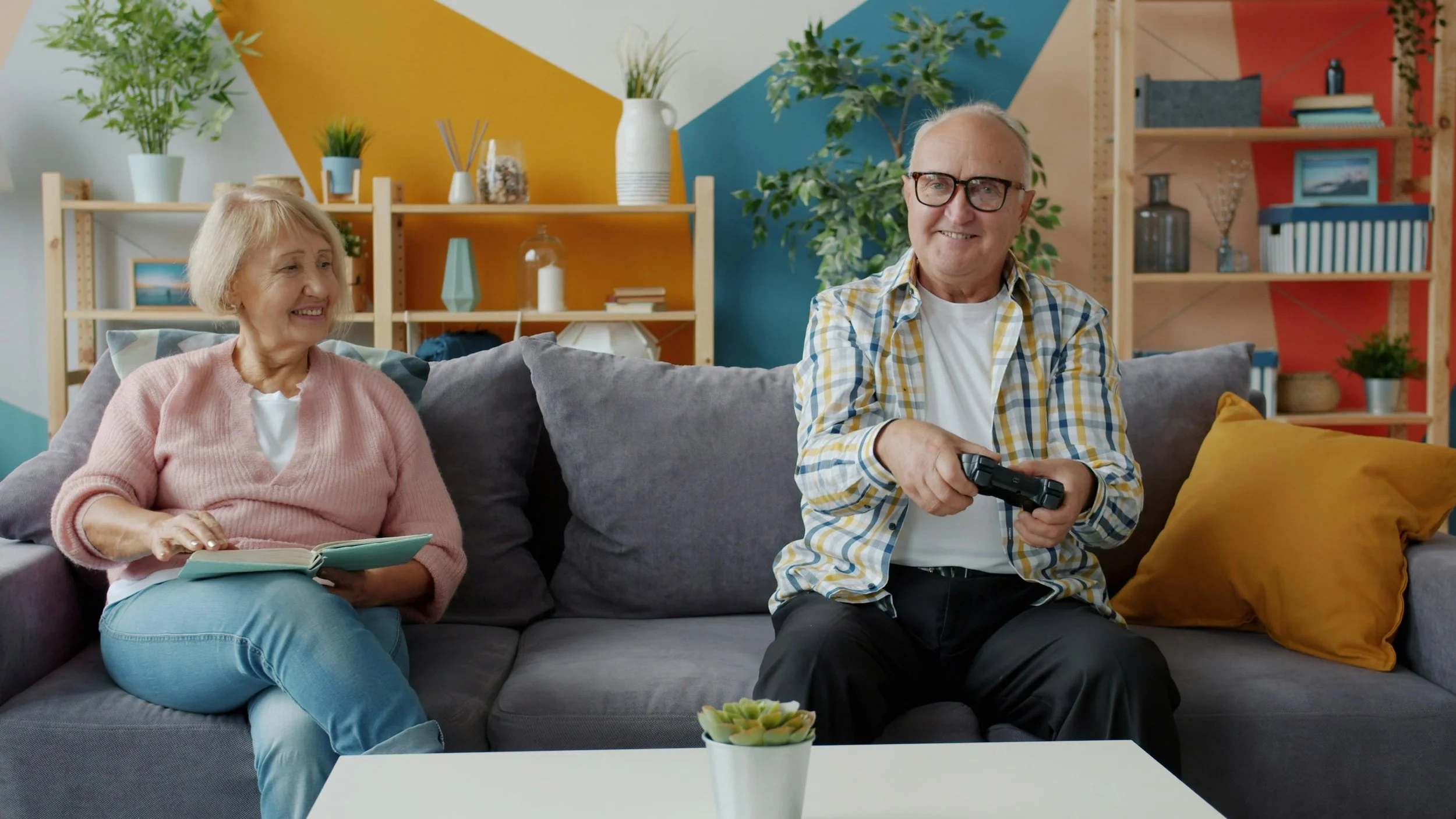 An elderly woman and man are sitting on a gray couch in a colorful living room. The woman is holding a book and smiling, while the man is holding a video game controller and smiling. The room has a modern, vibrant design with decorative shelves, plants, and colorful accent walls.