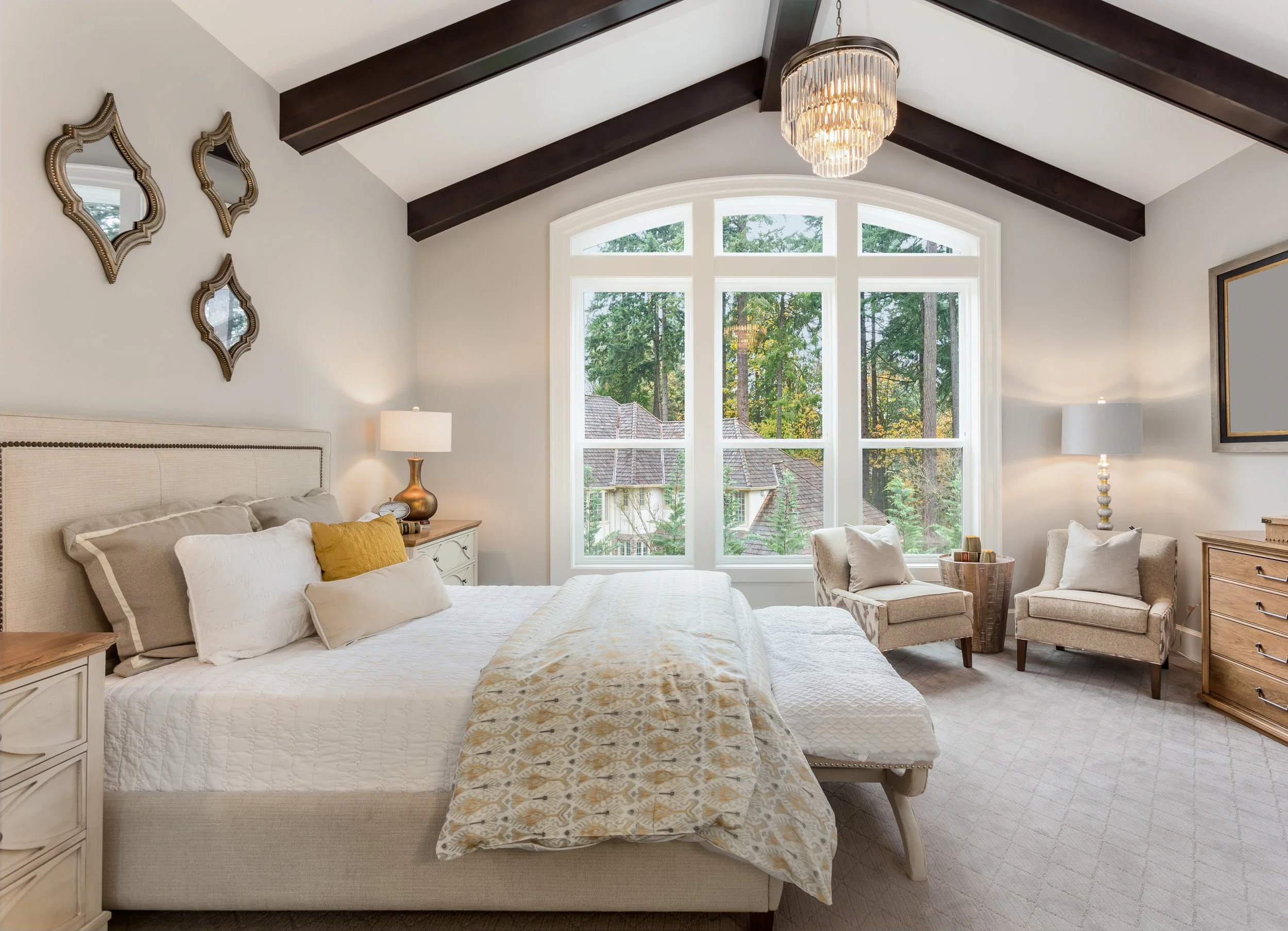 Bright bedroom with large arched window showing trees outside, beige upholstered bed with white bedding and pillows, two beige armchairs with throw pillows, wooden side table, and a dresser, decorated with a chandelier, mirrors, and lamps.