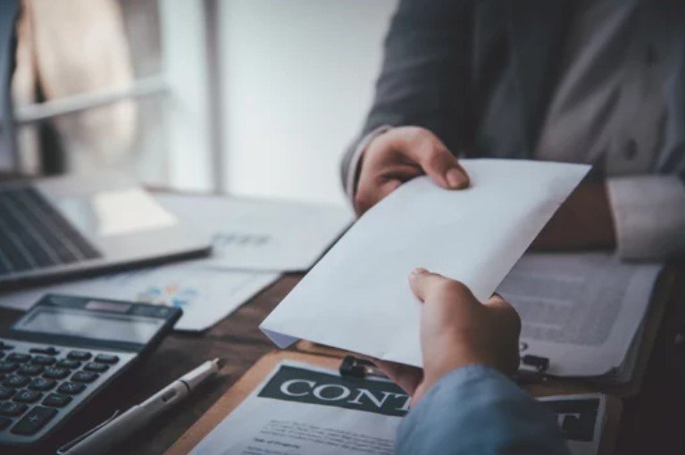 Person handing over a white paper to another person at a desk with a laptop, calculator, and financial documents.