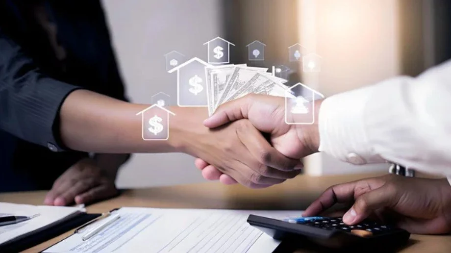 Two people shaking hands over a table with documents and a calculator, with a digital overlay of dollar signs and house icons representing real estate or financial exchange.