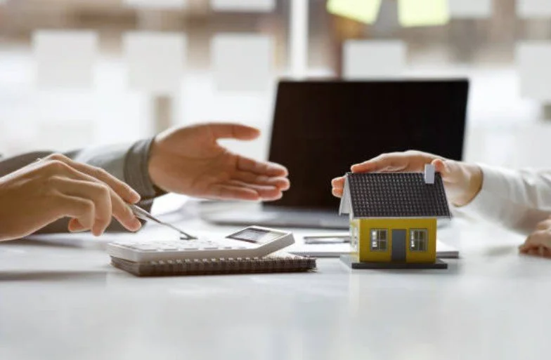 Two people discussing a real estate transaction at a table, with a small house model and a laptop present.