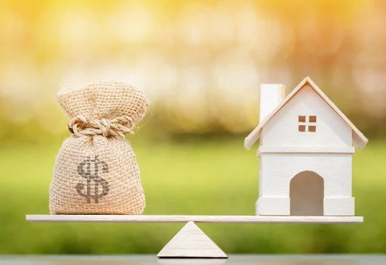 A small white house model balanced on a seesaw against a blurred outdoor background, with a burlap sack decorated with a dollar sign on the opposite side.