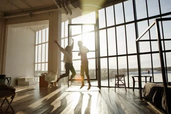 Two people jumping and dancing inside a bright, spacious room with large windows showing an outdoor view.