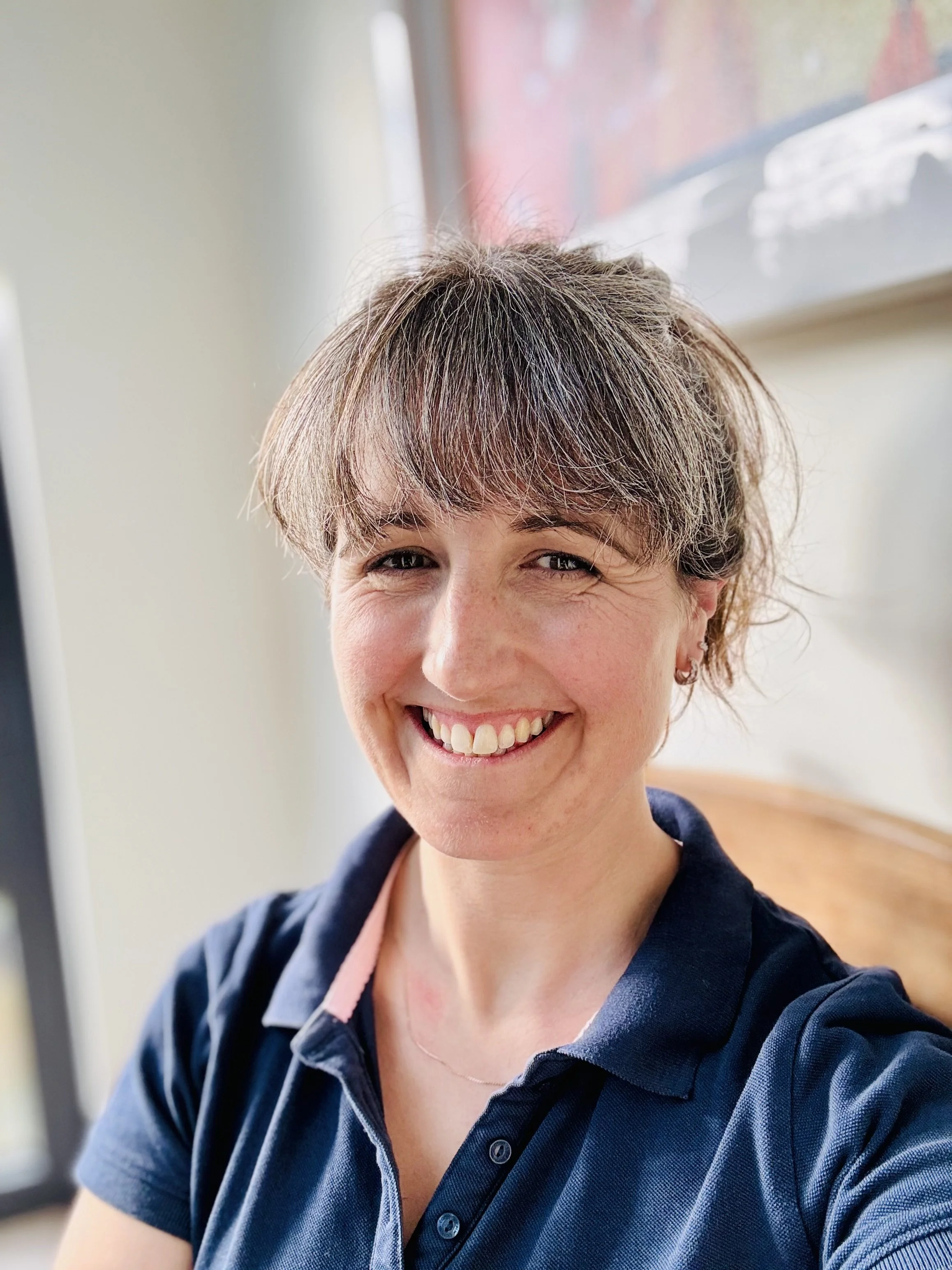 Close-up of a smiling woman with short, layered brown hair wearing a navy blue polo shirt, indoors with blurred background.