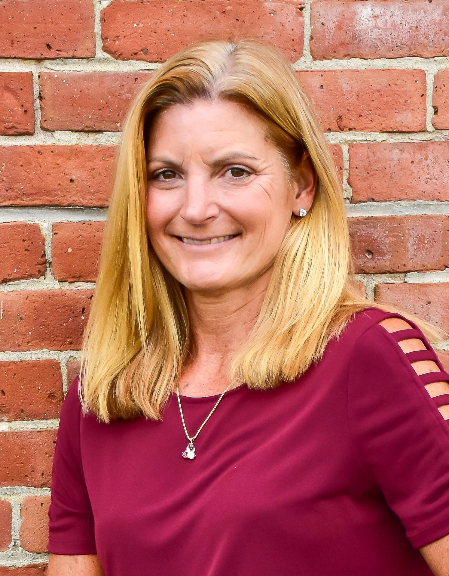 A woman with shoulder-length blonde hair, smiling, standing in front of a red brick wall, wearing a burgundy top with cutout shoulder details and a silver necklace.