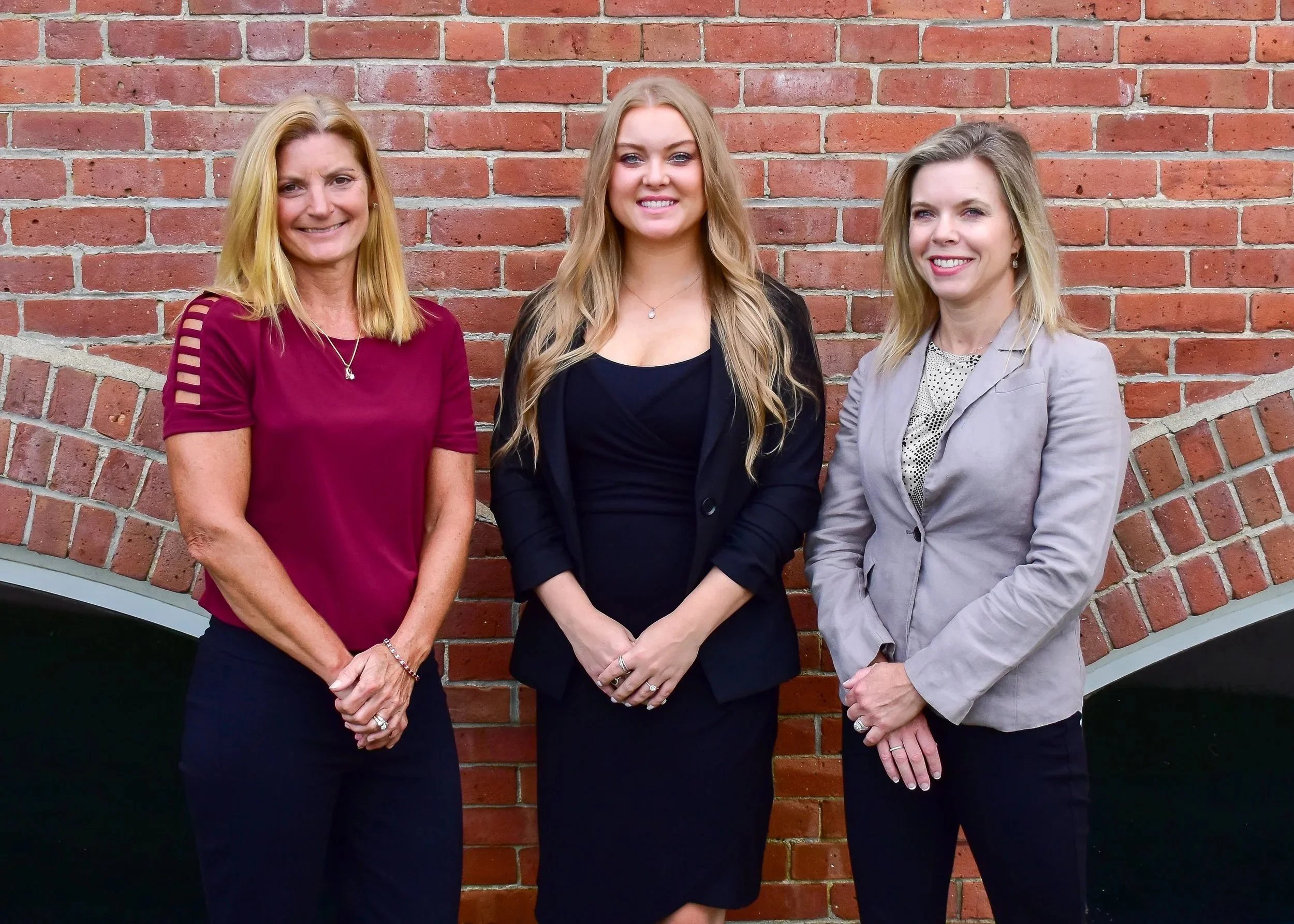Three women standing in front of a brick wall, smiling, dressed in business casual attire.