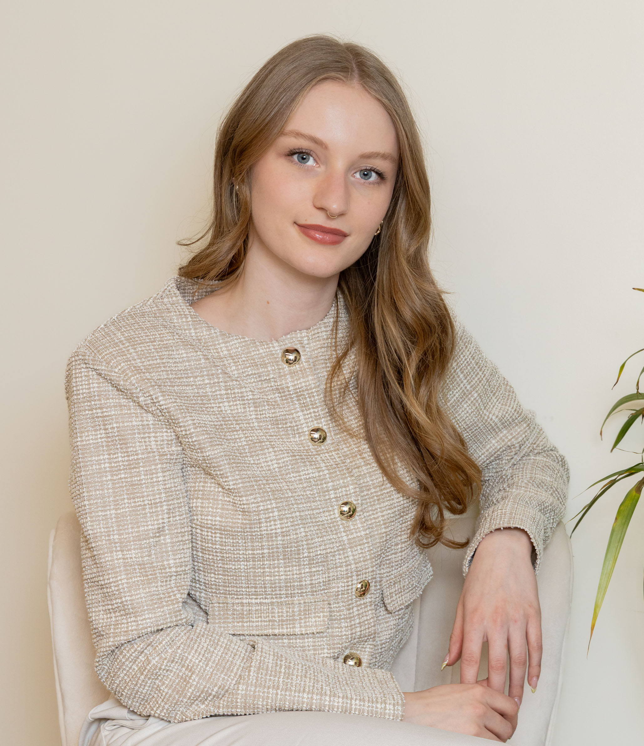 A young woman with long, wavy red hair and blue eyes, sitting on a beige chair against a neutral background, wearing a beige textured jacket with large decorative buttons and cream-colored pants.