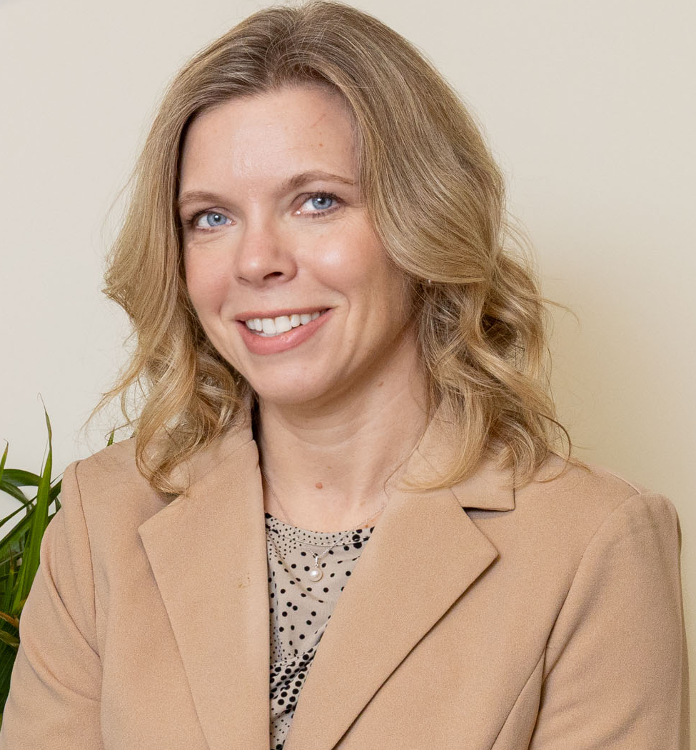 A portrait of a smiling woman with blonde, wavy hair wearing a beige blazer and a polka dot blouse, sitting in front of a plain light-colored wall with a green plant partially visible on her left.
