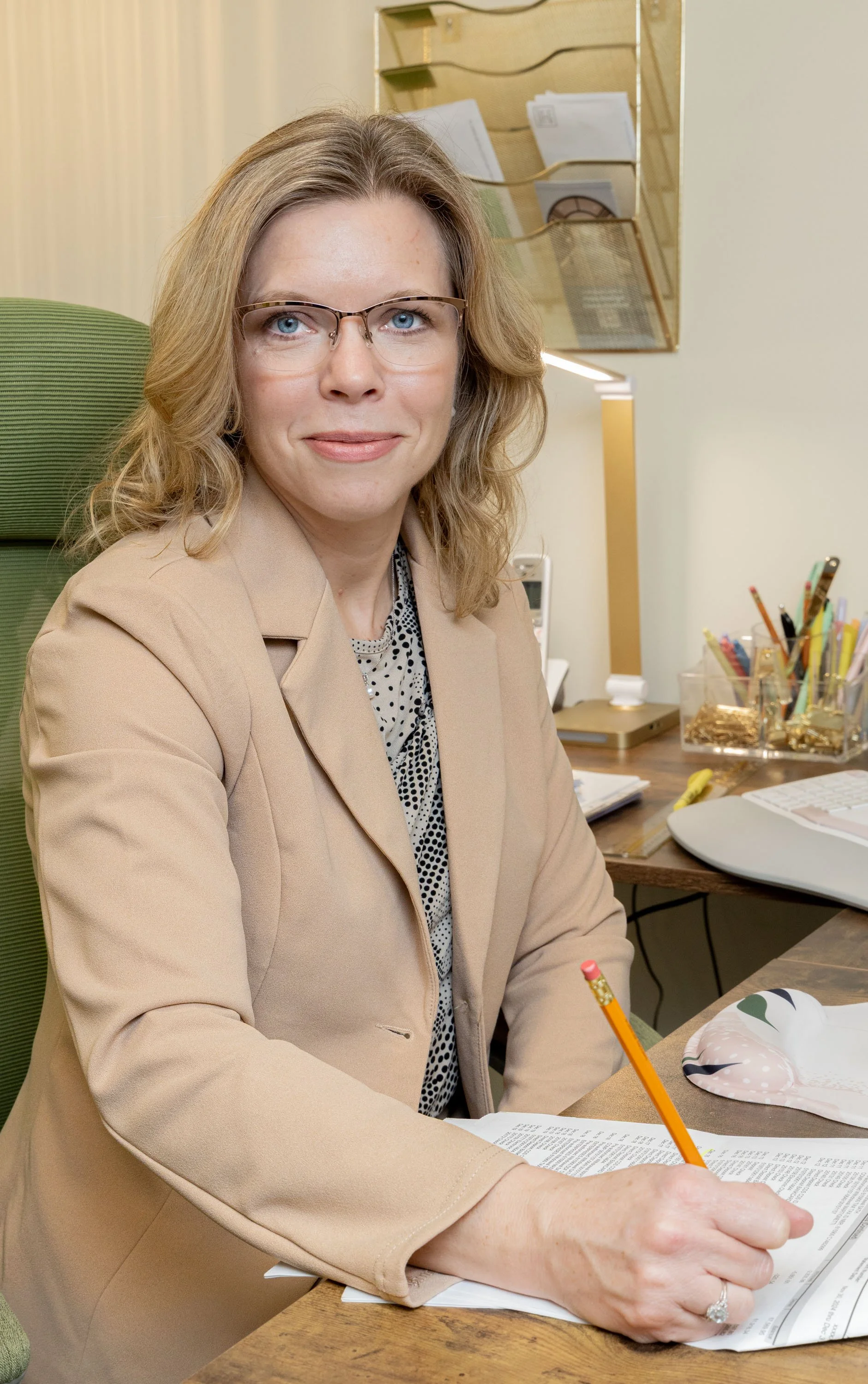 A woman with blonde hair and glasses sitting at a desk, writing with a pencil on paper, in an office setting.