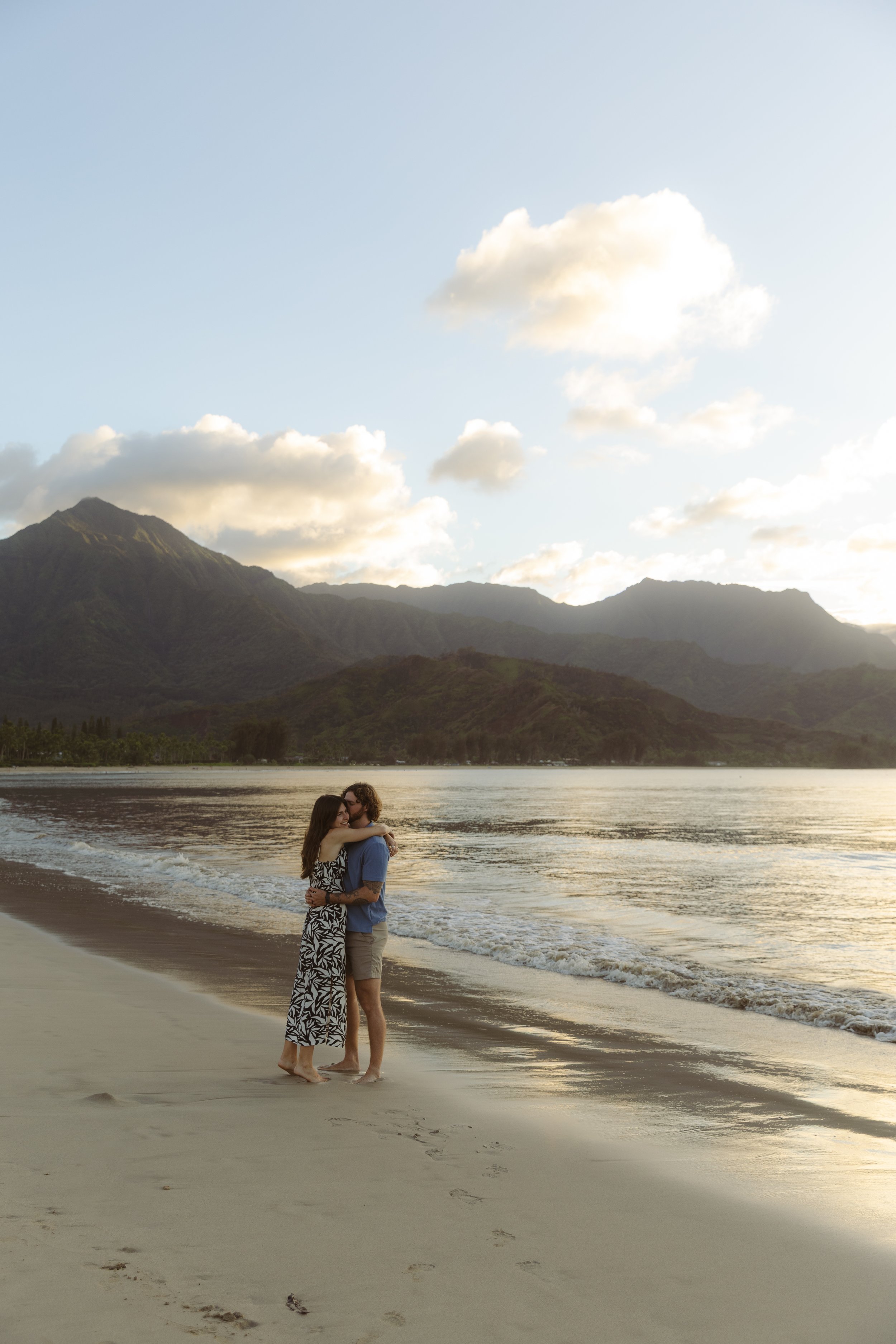 A couple hugging on a sandy beach with mountains and a cloudy sky in the background during sunset.