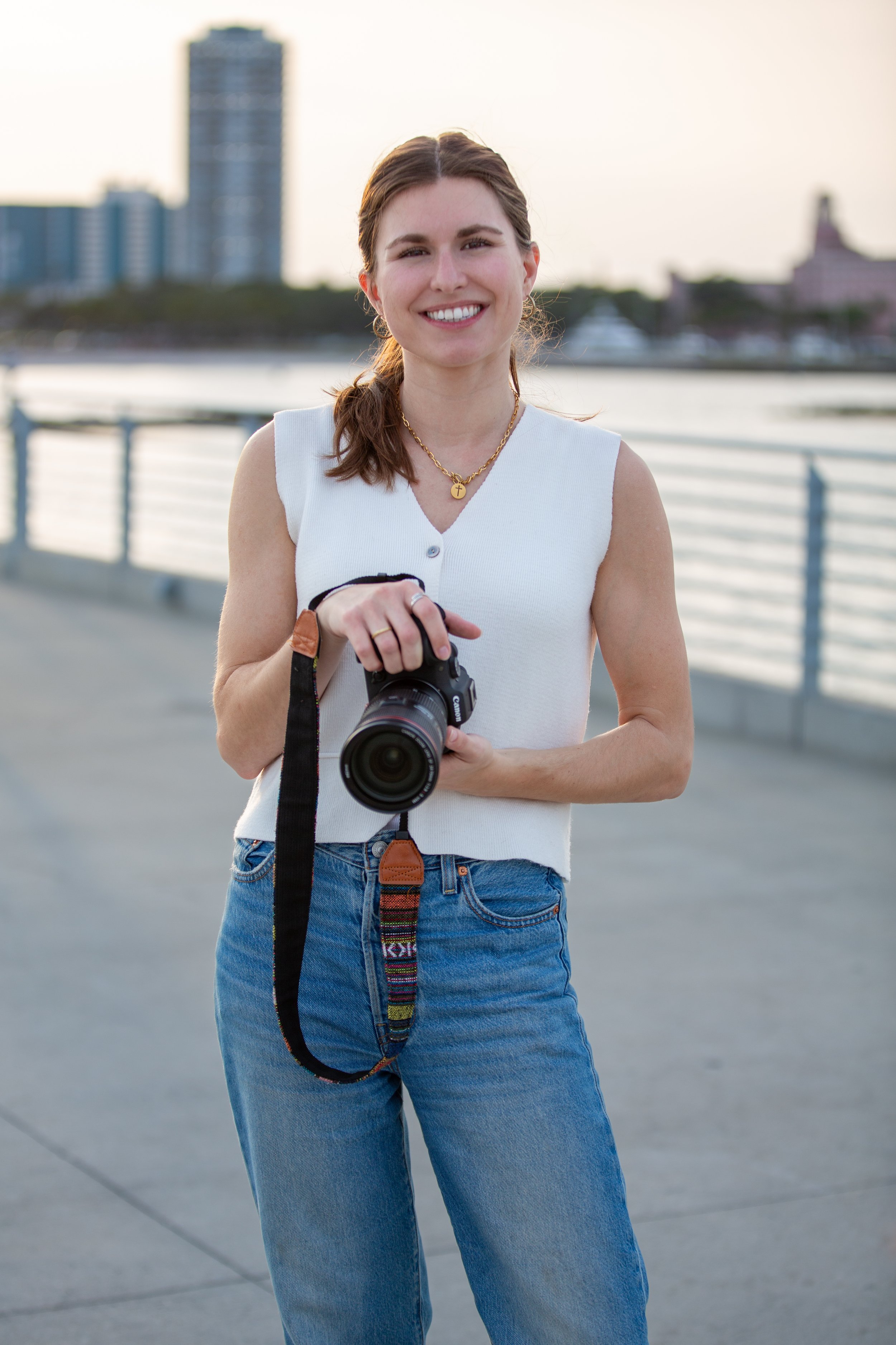 A young woman smiling, holding a camera, standing outdoors by a waterfront with city buildings in the background.