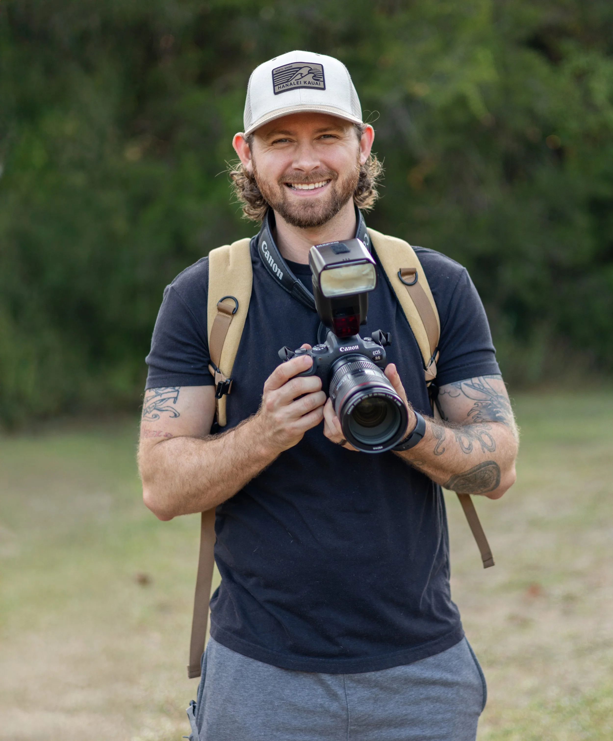 A smiling man with a beard, wearing a beige cap, black t-shirt, gray shorts, and a backpack, holding a Canon camera with a flash mounted on top, standing outdoors in a grassy area with trees in the background.