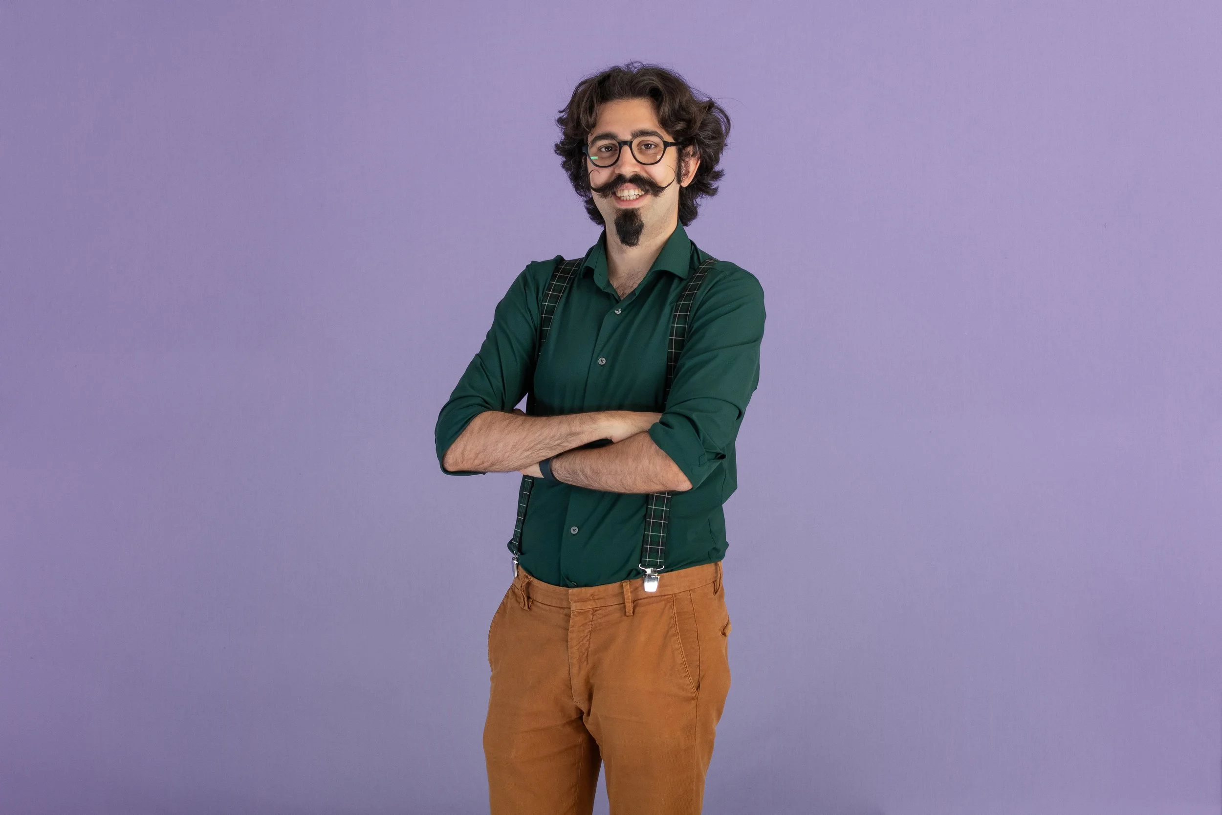 Gianmarco Pallaoro, uomo con capelli riccioli e barba, indossa occhiali, camicia verde con i risvolti arancioni, pantaloni marroni, con bretelle e sorriso, su sfondo viola.