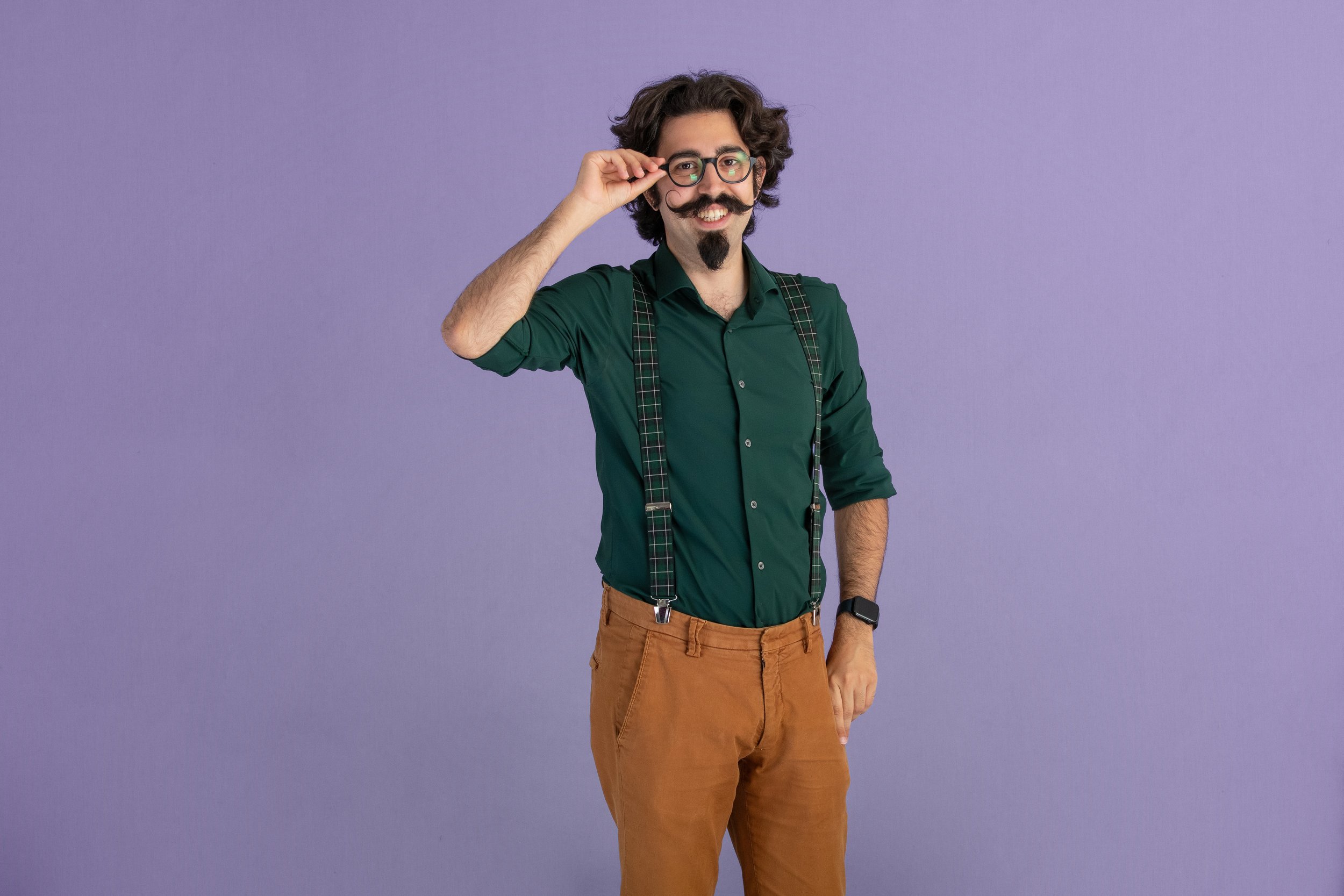 Gianmarco Pallaoro, uomo con occhiali, baffi e barba corta, vestito con camicia verde, pantaoni marroni, con bretelle, che sorride e si regola gli occhiali davanti a uno sfondo violetta.