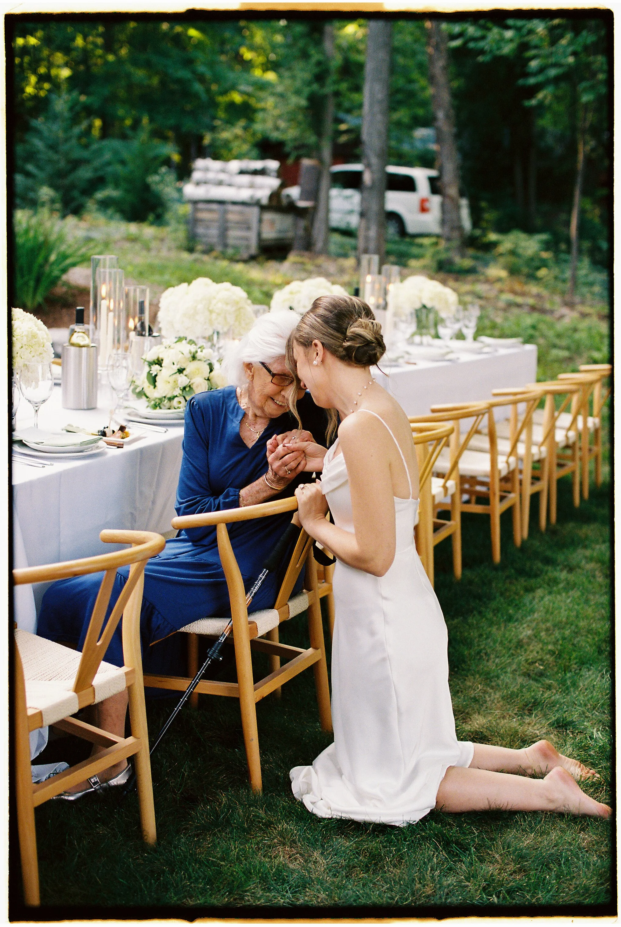 A bride kneeling on grass, holding hands with an elderly woman seated at a decorated outdoor wedding reception table, both smiling and touching foreheads.