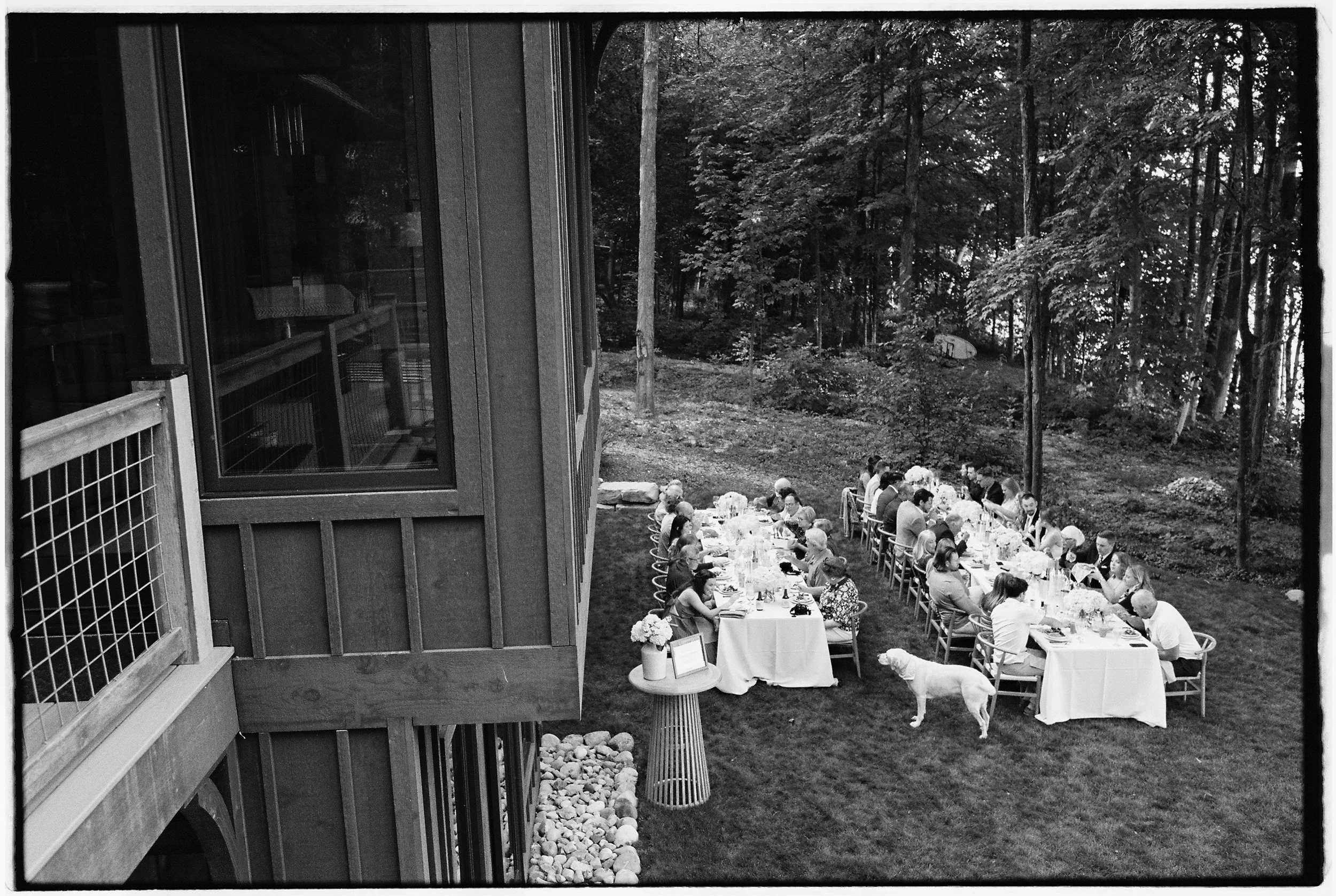 Outdoor dinner party with multiple tables set for guests, surrounded by a wooded area.