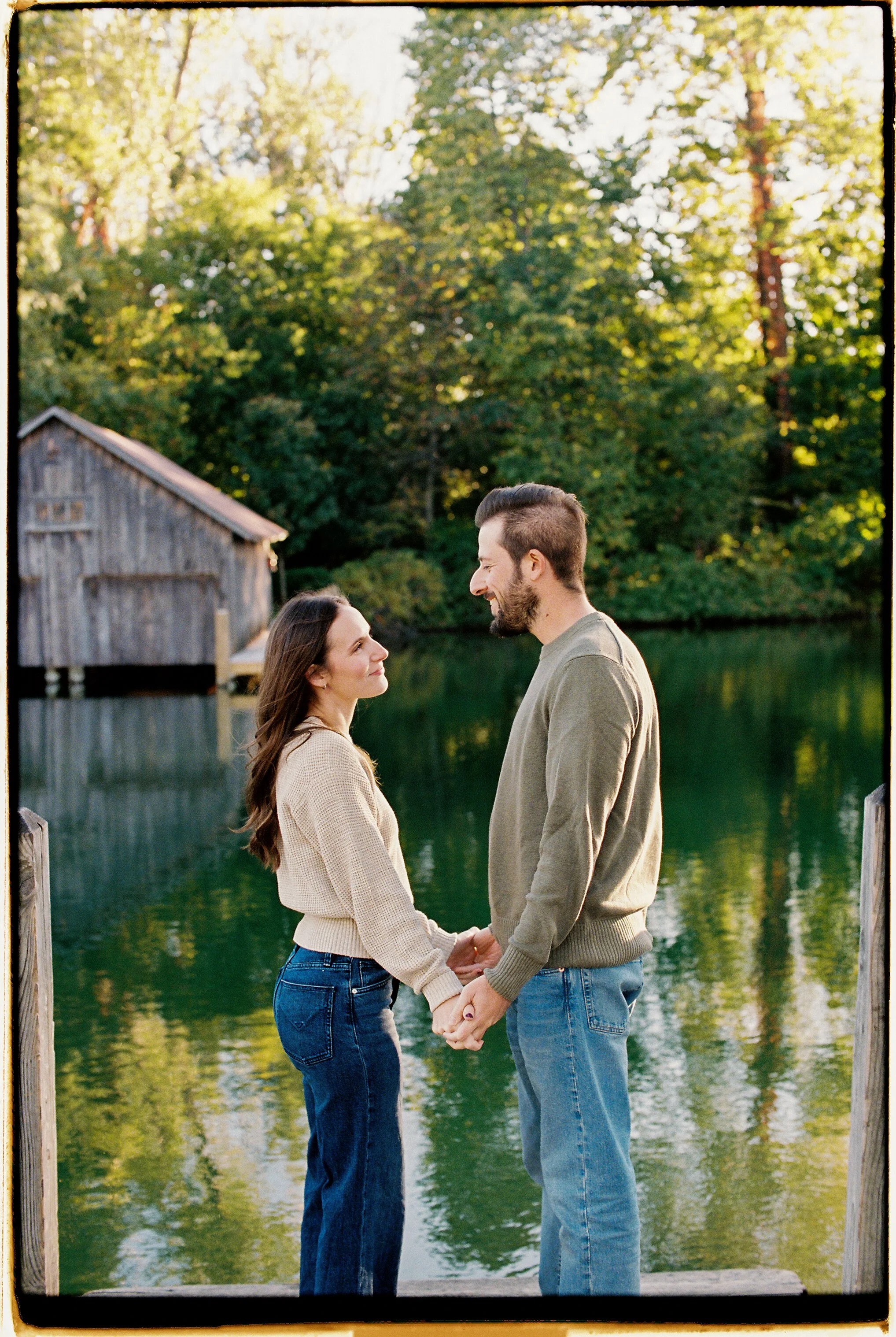 A young couple holding hands and gazing at each other on a dock near a lake during sunset, with a wooden boathouse and green trees in the background.