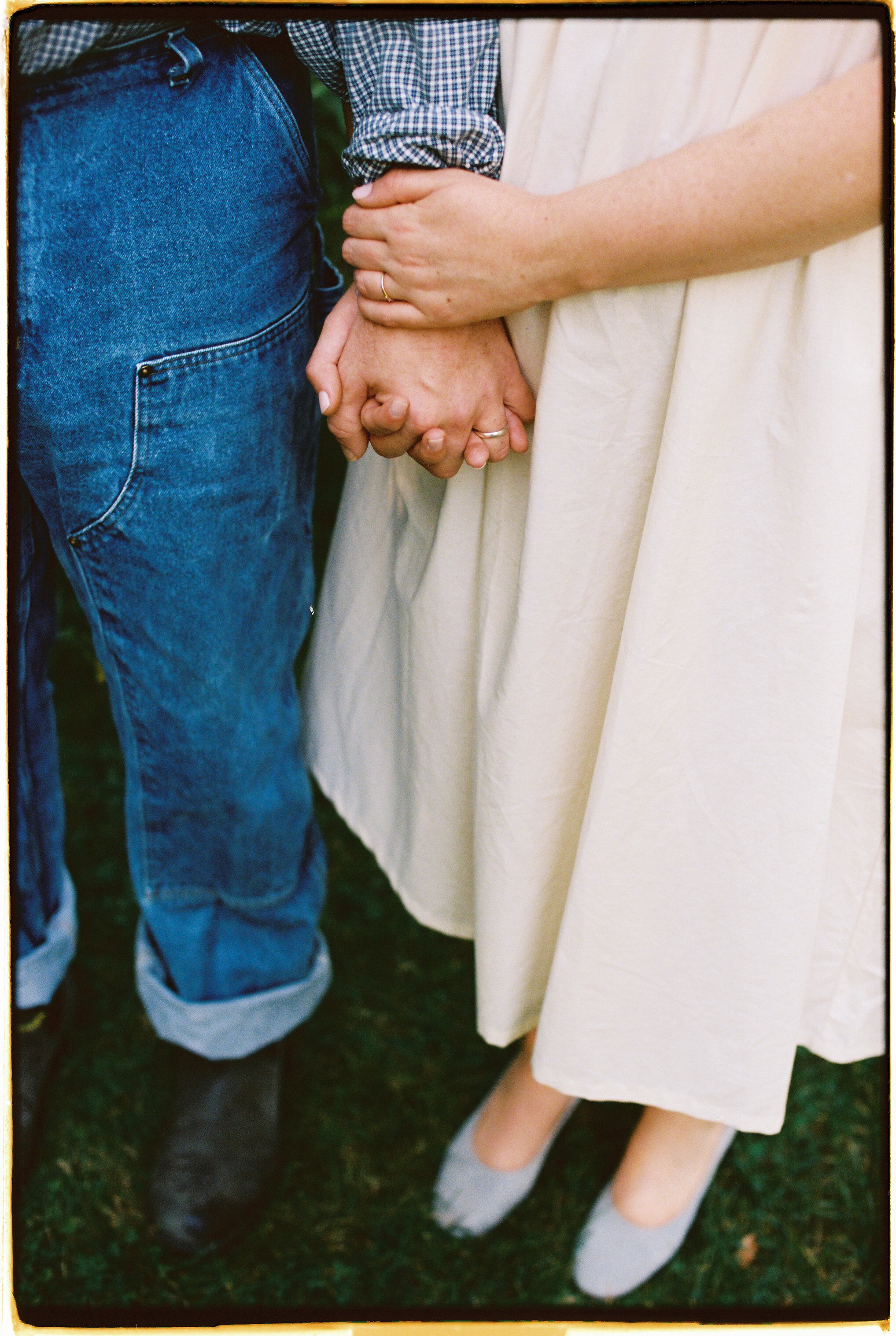 Close-up of a couple holding hands, with the man wearing blue jeans and a checkered shirt, and the woman in a cream dress and gray shoes.