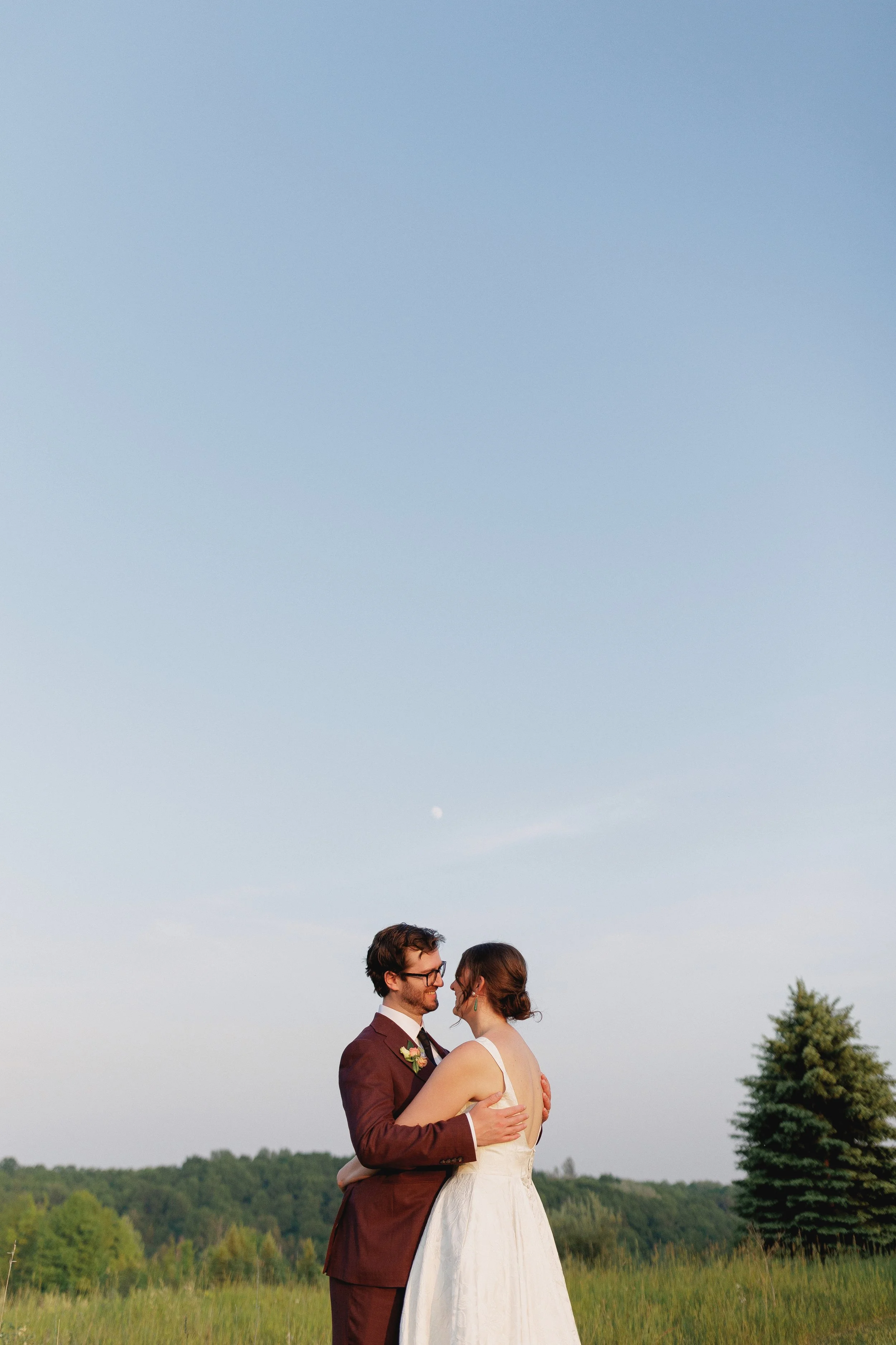A newlywed couple with the groom in a dark suit and the bride in a white dress, standing in a green field, embracing and looking into each other's eyes with a clear blue sky and a visible moon in the background.