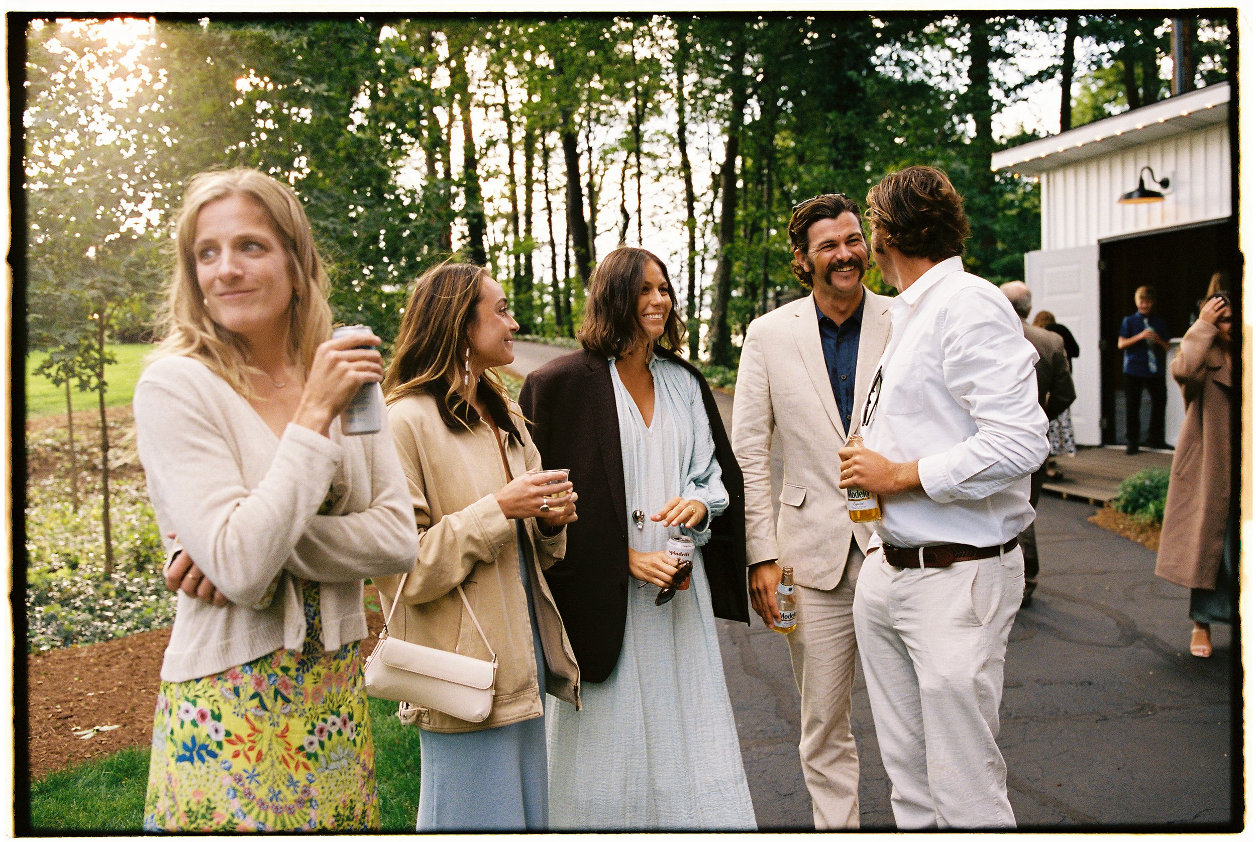 Group of friends socializing outdoors at a gathering, with trees and a shed in the background, some holding drinks and engaging in conversation.