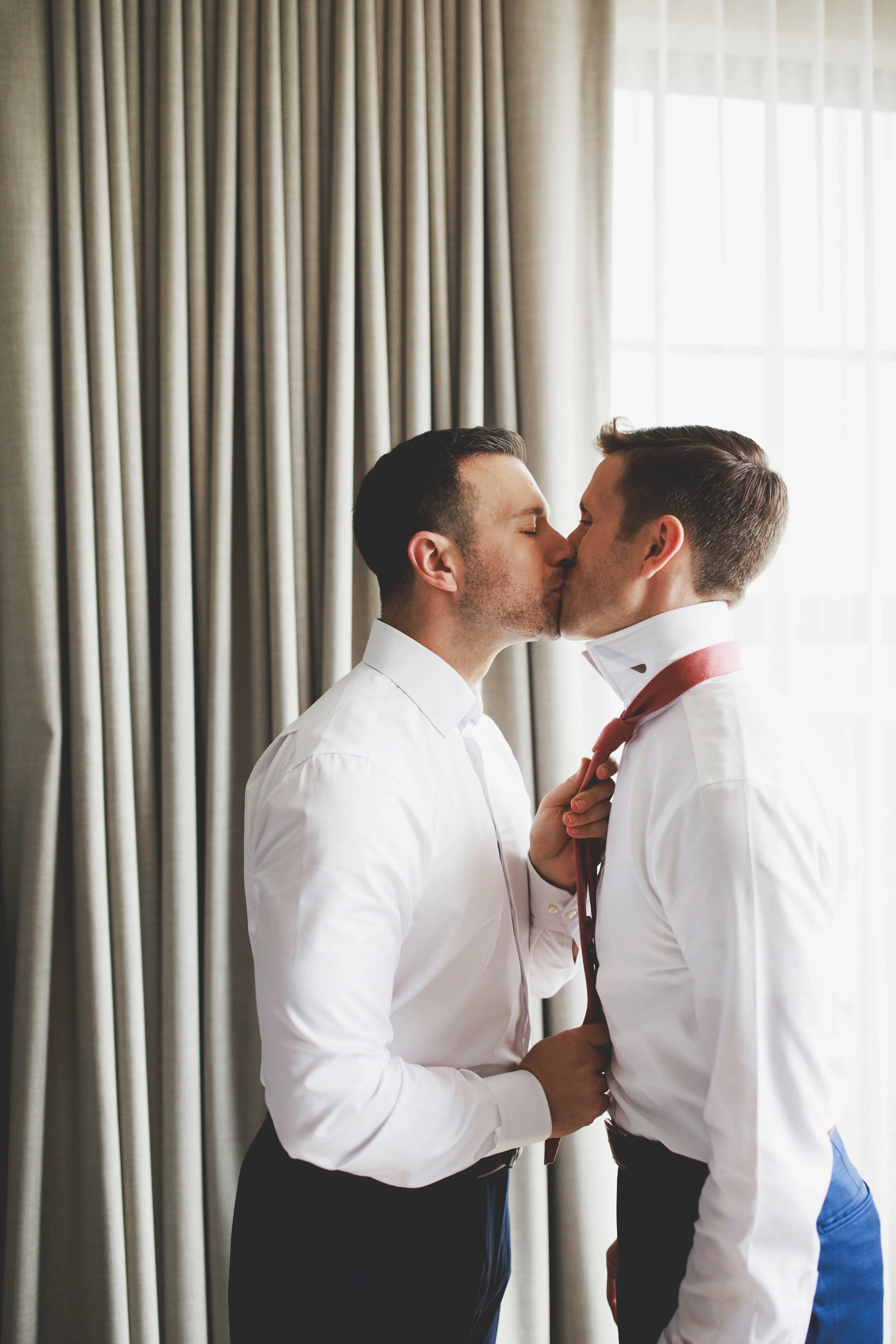 Two men dressed in white shirts are sharing a kiss, with one adjusting the other's red tie, in front of beige curtains and a window in the background.