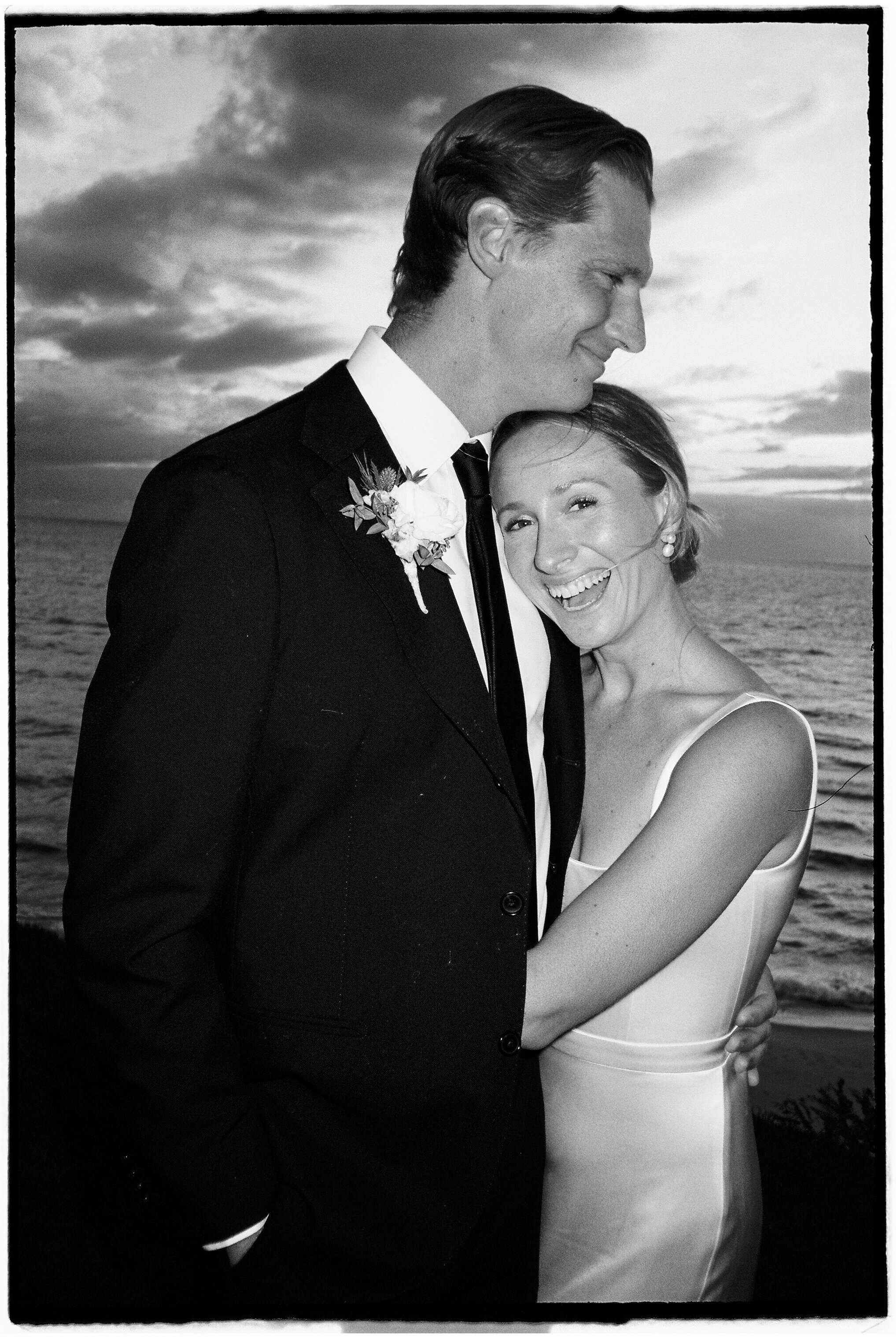 A Black and white photo of a happy wedding couple embracing near the ocean at sunset, the groom in a suit with a boutonniere, and the bride in a satin dress smiling.