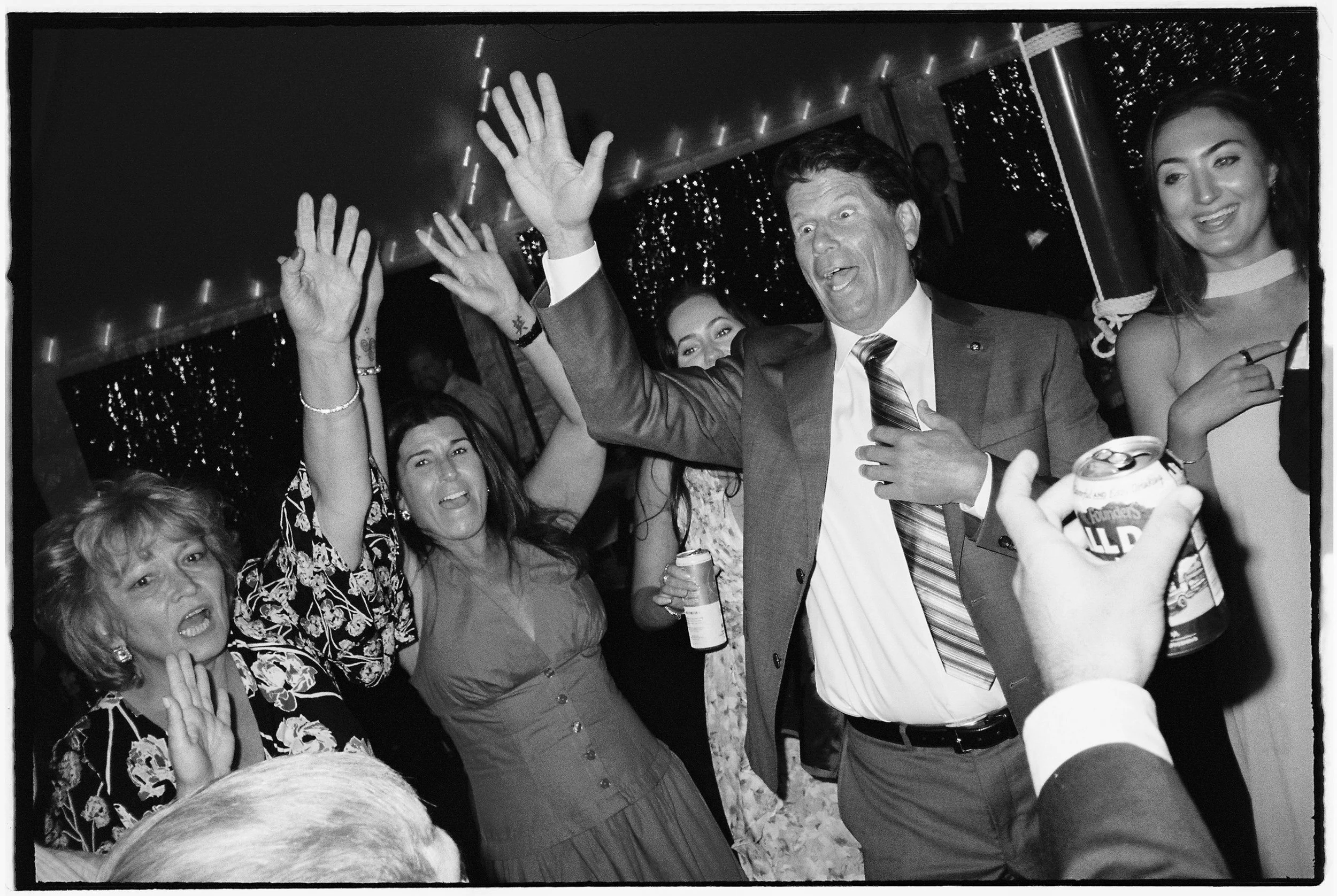 People celebrating at a party, raising hands, holding drinks, and smiling, with festive lights in the background.