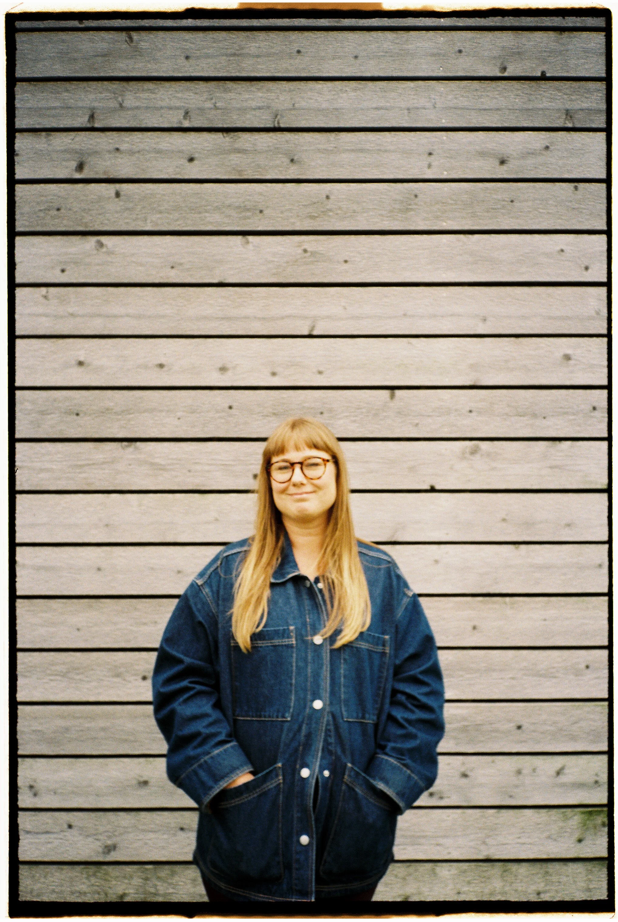 A woman with long blonde hair, glasses, and a denim jacket standing in front of a wooden wall, smiling with hands in pockets.