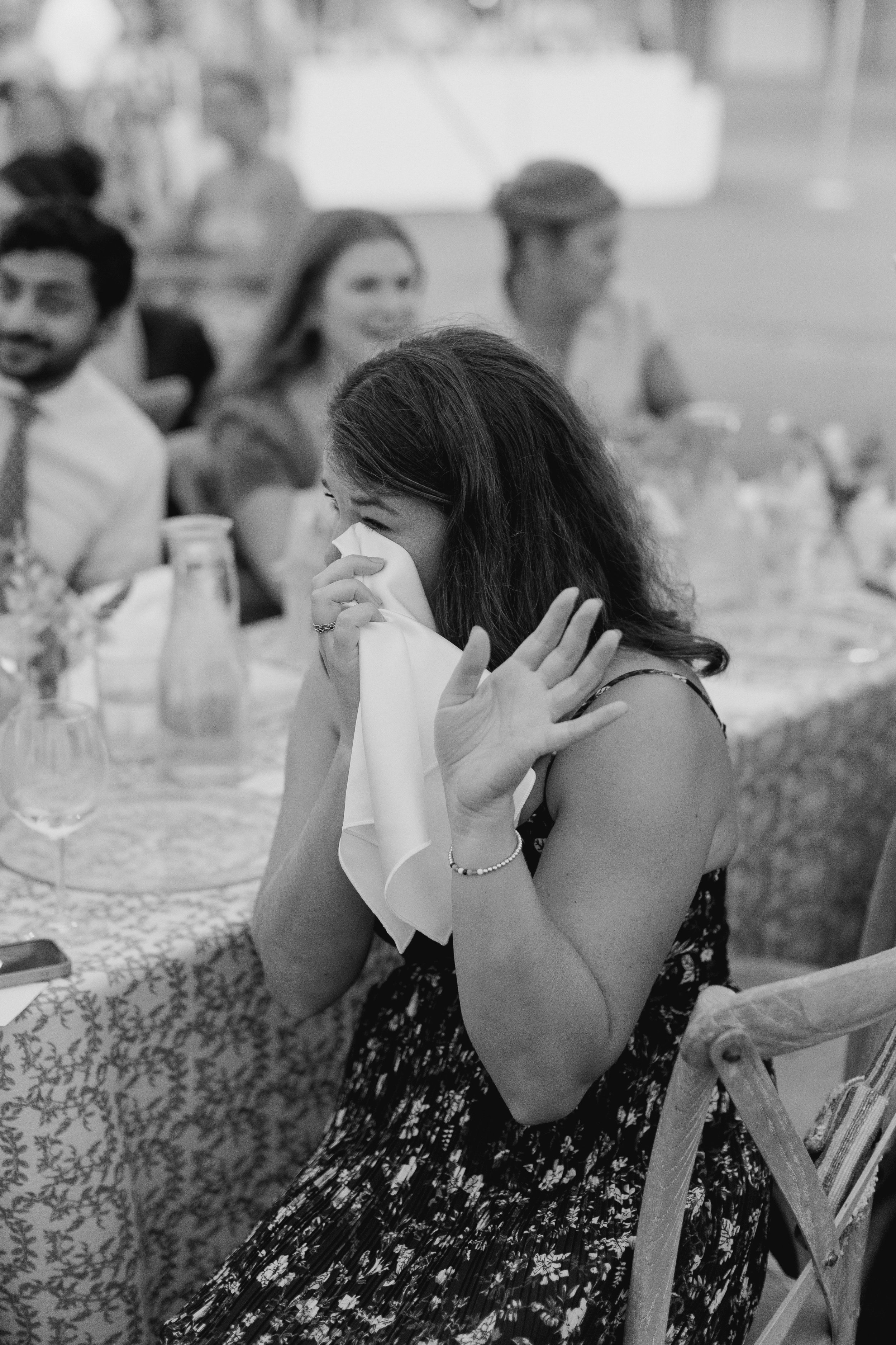 A woman sitting at a dinner table, wiping away tears with a napkin, surrounded by friends or family at a celebration or gathering.