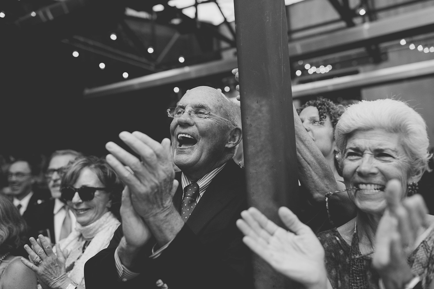 Group of seniors clapping and smiling at an indoor event, with some wearing sunglasses, in black and white.