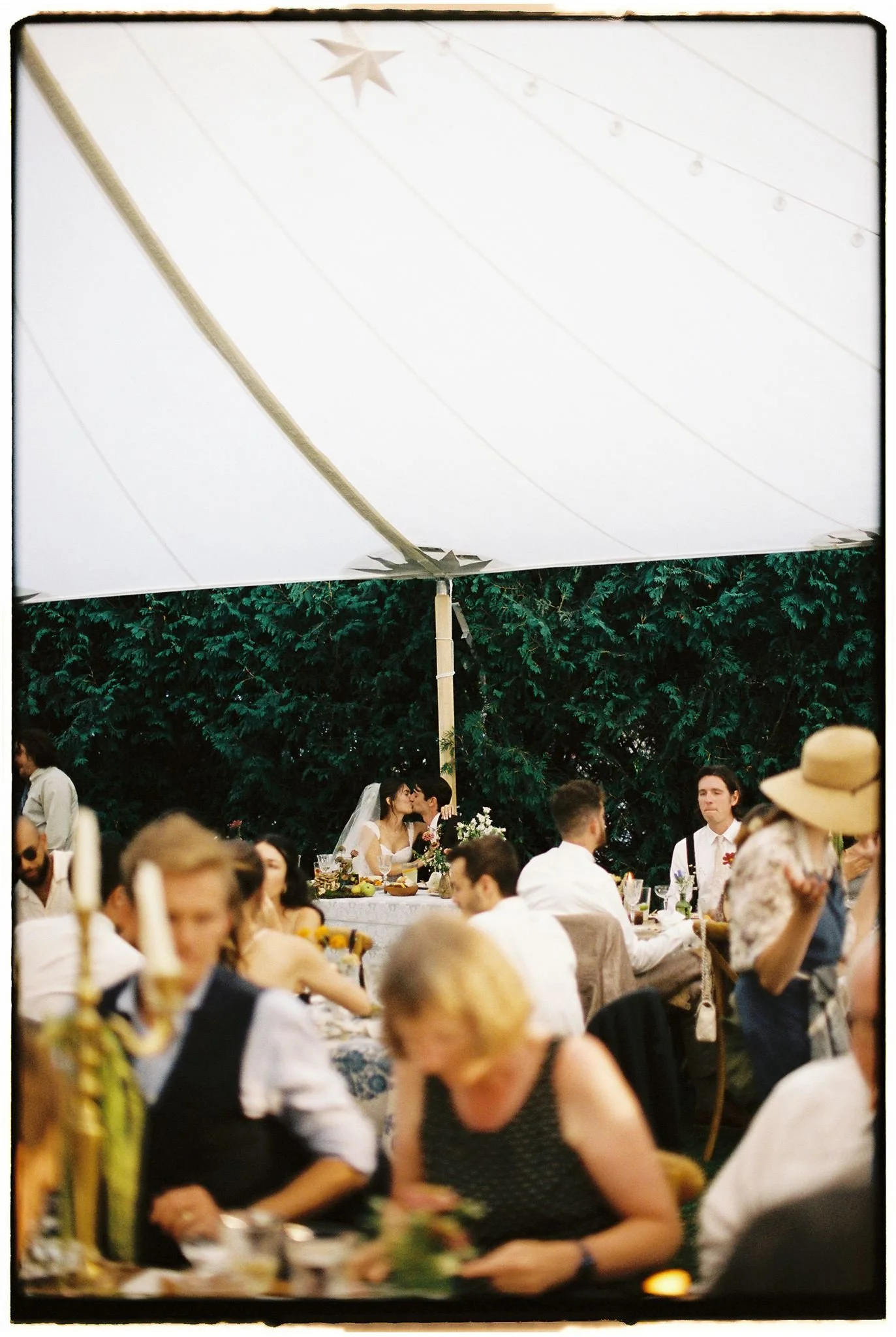 A wedding reception under a large white tent with hanging stars and string lights. The bride and groom are kissing at a head table decorated with flowers. Guests are seated at tables, some engaged in conversation or looking at their phones.