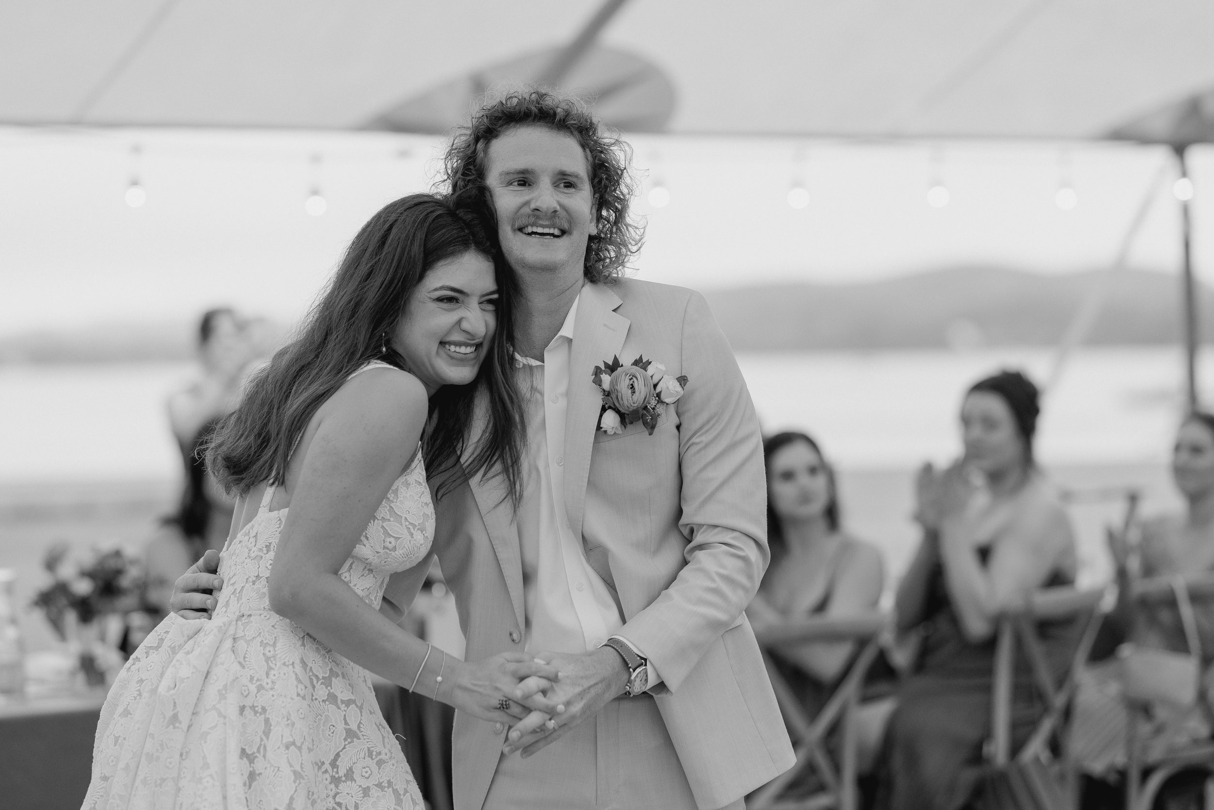 A black and white photo of a bride and groom dancing at their wedding reception outdoors, with guests seated at tables in the background.