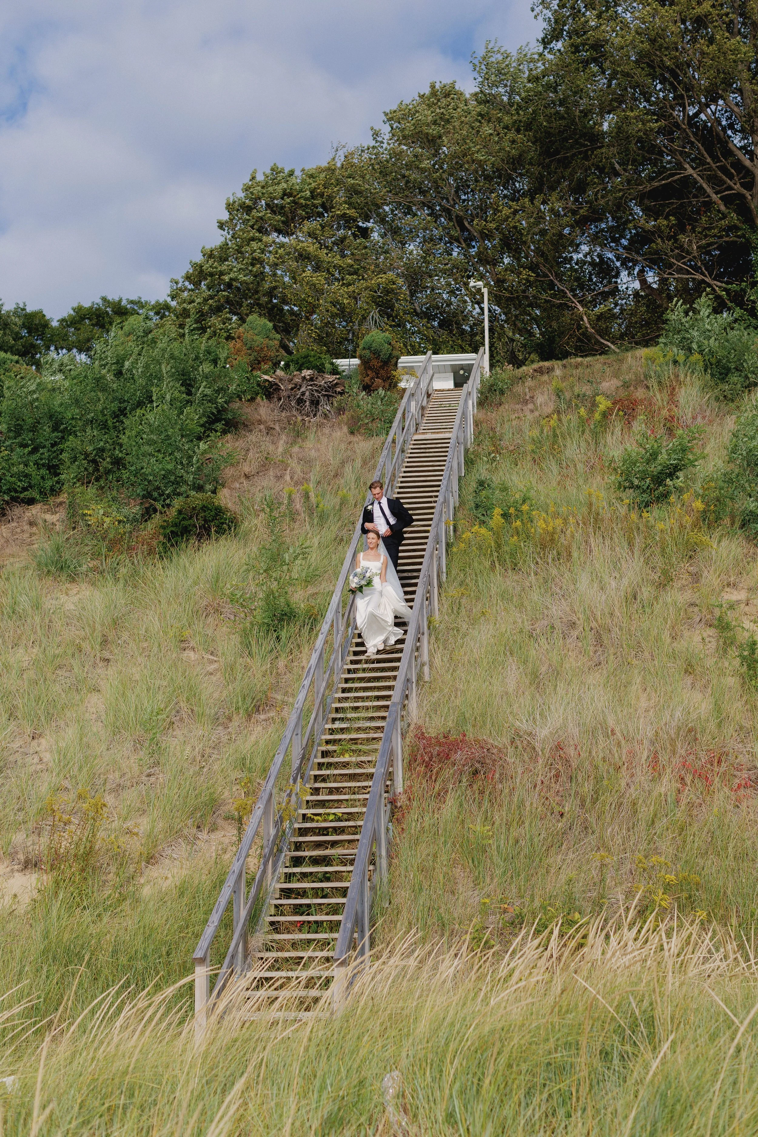 A bride and groom descending a long staircase outdoors on a grassy hillside with trees in the background.