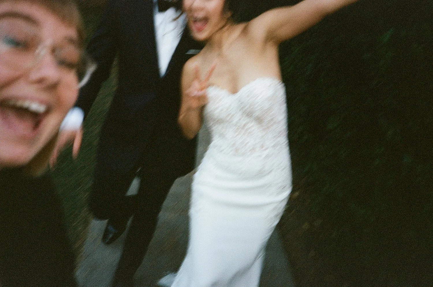 Three people having fun, with one woman in a white strapless dress making a peace sign and two men in suits smiling and posing for a selfie.
