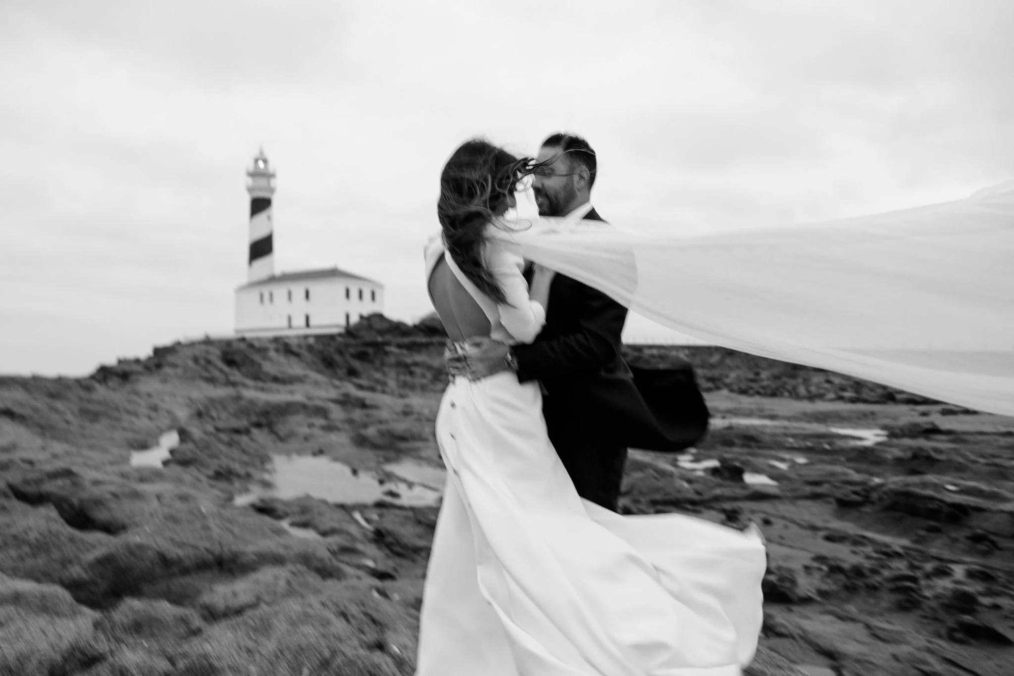 Pareja de novios bailando junto al mar con un faro al fondo, fotografía de boda en blanco y negro.