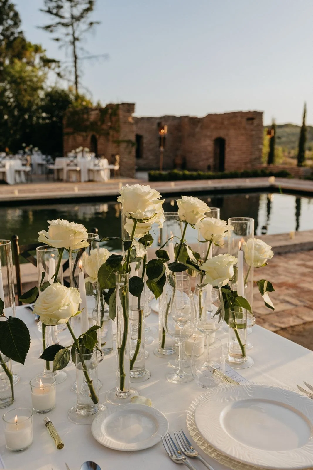 Mesa de boda al aire libre decorada con rosas blancas y velas, junto a unas ruinas históricas y naturaleza.