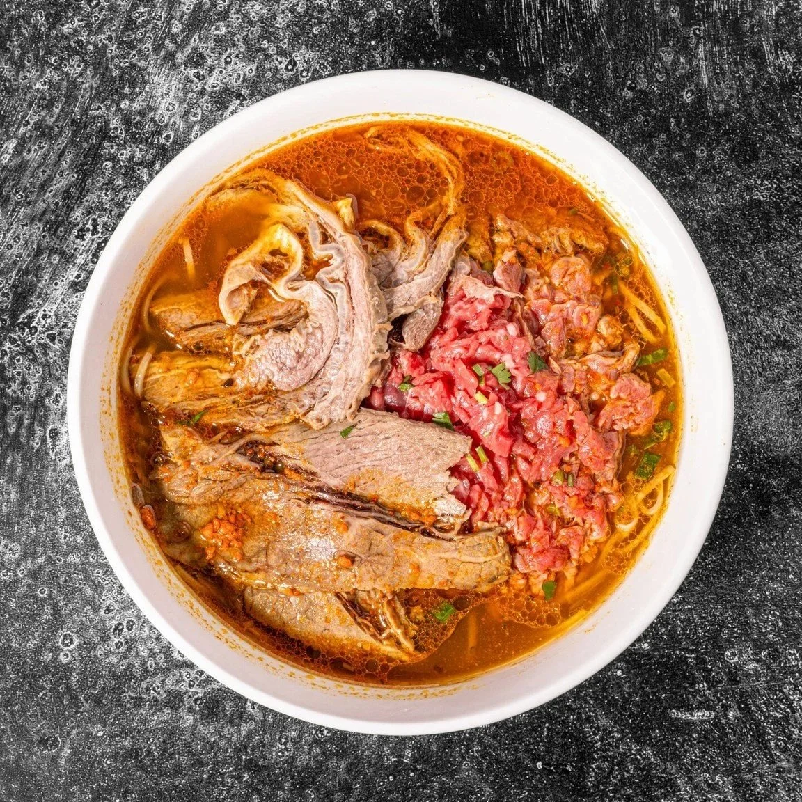 A bowl of ramen soup topped with slices of beef and diced tomatoes on a dark textured surface.