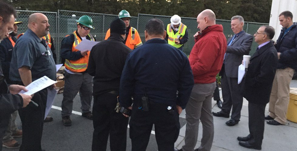 Group of construction and business professionals standing and listening during an outdoor meeting, some holding documents.