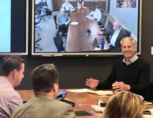 A business meeting with several people in a conference room, including a man with gray hair and a black sweater sitting at a large wooden table, with others listening. A large screen behind him shows a video call with additional participants seated at a similar table.