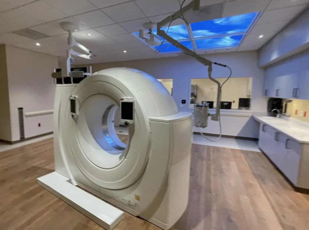 CT scanner in a modern medical imaging room with overhead equipment, counters, and wood-look flooring.