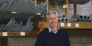 A smiling man stands in a warehouse aisle with shelves of gray pipes behind him.