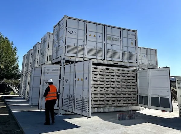 A worker in an orange safety vest and helmet standing near stacked refrigerated shipping containers outdoors on a sunny day.