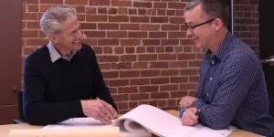 Two men smiling and shaking hands across a table outside, with a brick wall in the background.