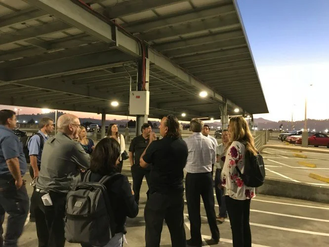 Group of people standing and talking in a parking lot near a building, with cars parked and a sunset in the background.