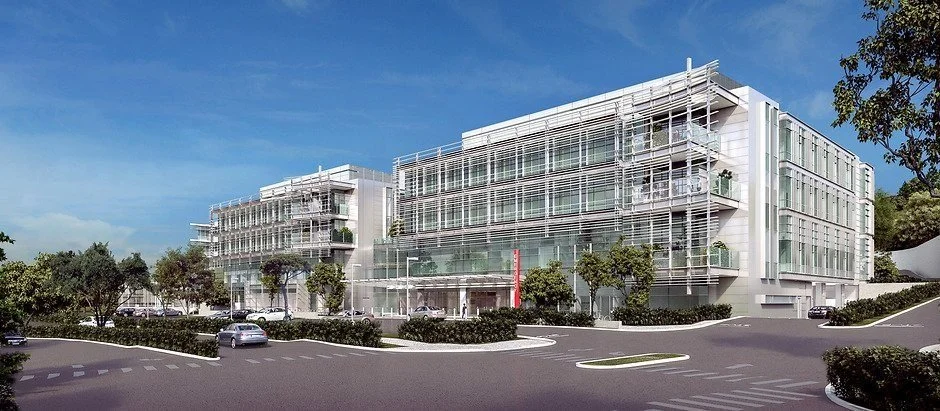 Modern multi-story medical office building with glass facade and white framing, surrounded by trees, parked cars, and a clear blue sky.