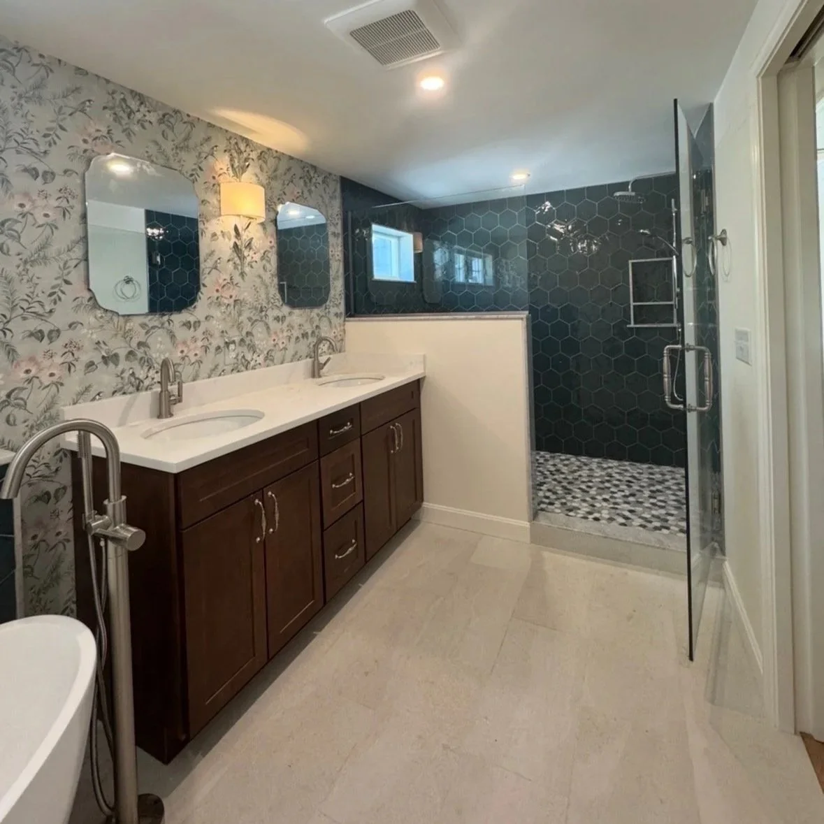 Modern bathroom with double vanity featuring dark wood cabinets and white countertop, three mirrors, floral wallpaper, a shower with black hexagonal tiles, and a small window. Built Structure Pro in Massachusetts