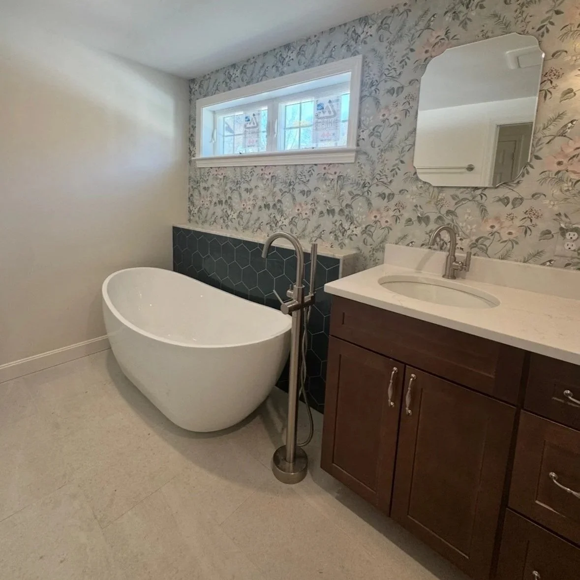 Modern bathroom with a white freestanding oval bathtub, a wooden vanity with a white countertop and sink, a rectangular mirror, a window, and floral wallpaper on one wall. Built Structure Pro in Massachusetts